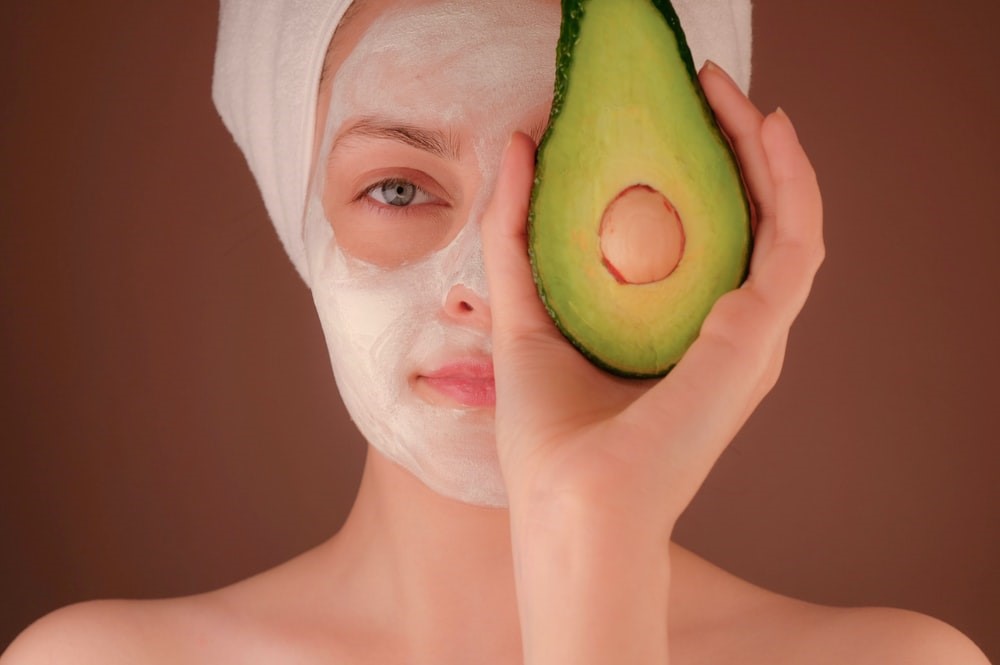 woman with facial holding an avocado to her face